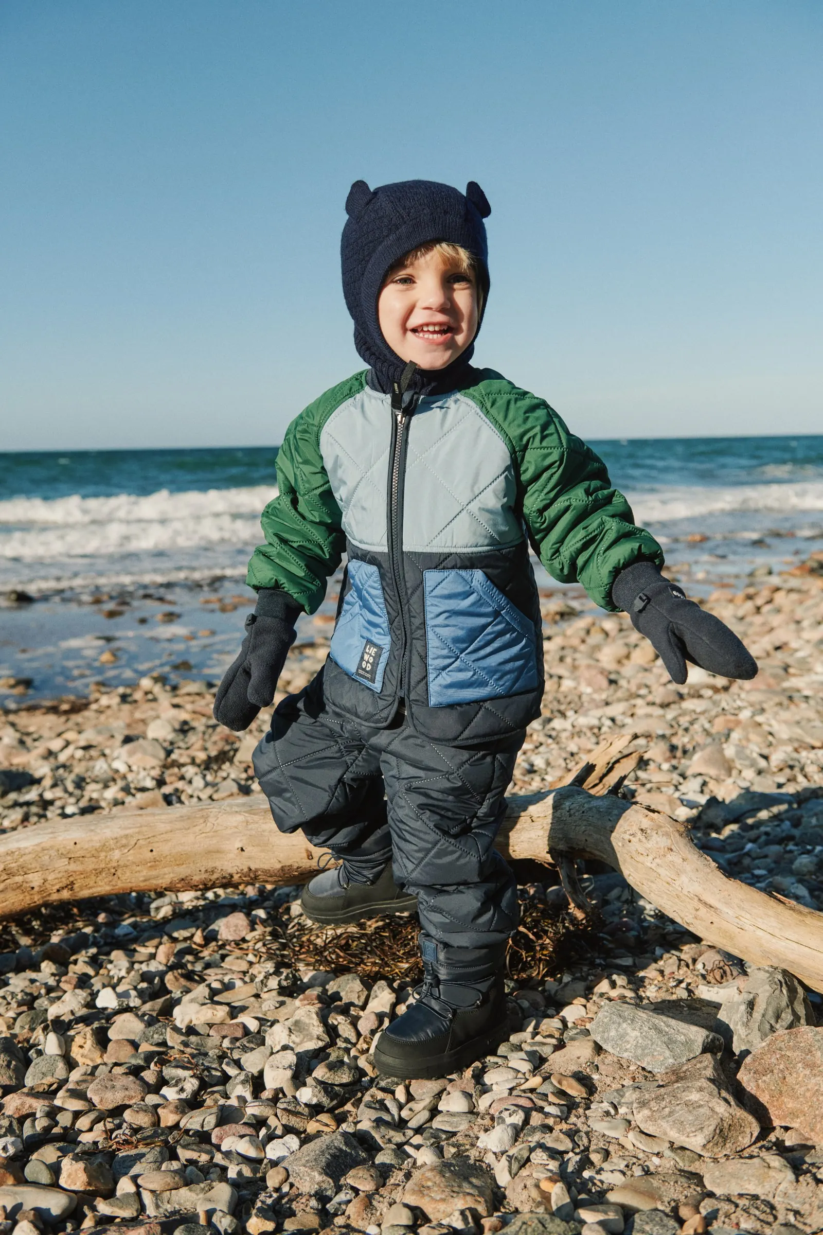 Kleinkind in gesteppter Winterjacke, Schneehose und Strickmütze mit Ohren steht lachend an einem Kiesstrand