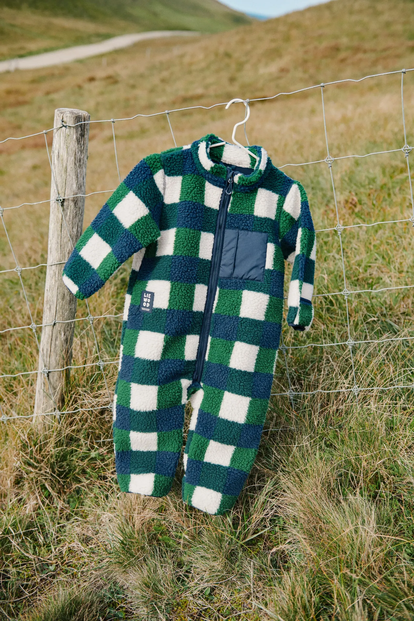 Kariertes Fleece-Overall für Kleinkinder hängt auf einem Bügel an einem Weidezaun