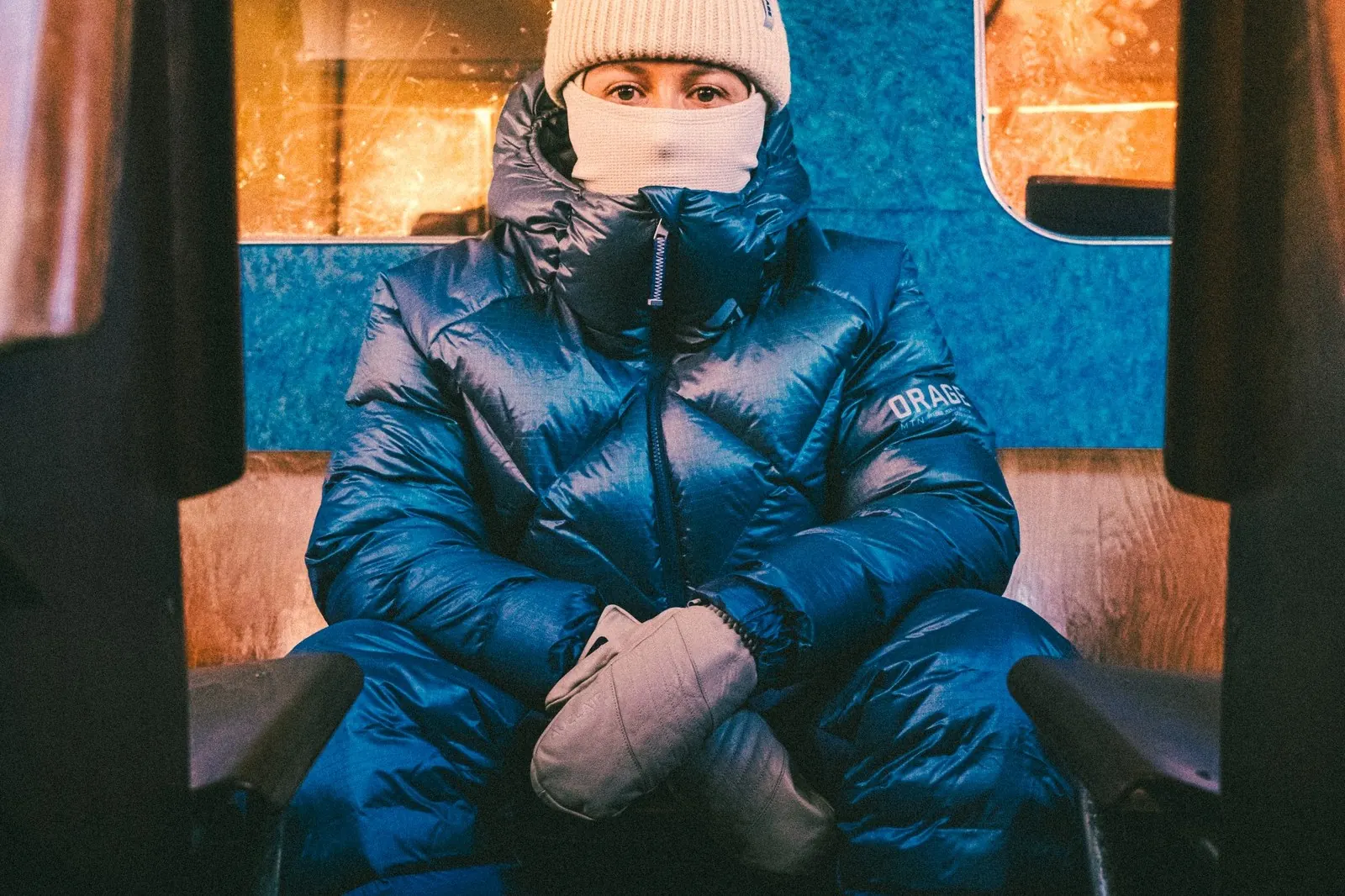 Person in a blue Orage Nimbus down jacket with a beanie and mittens in a train compartment