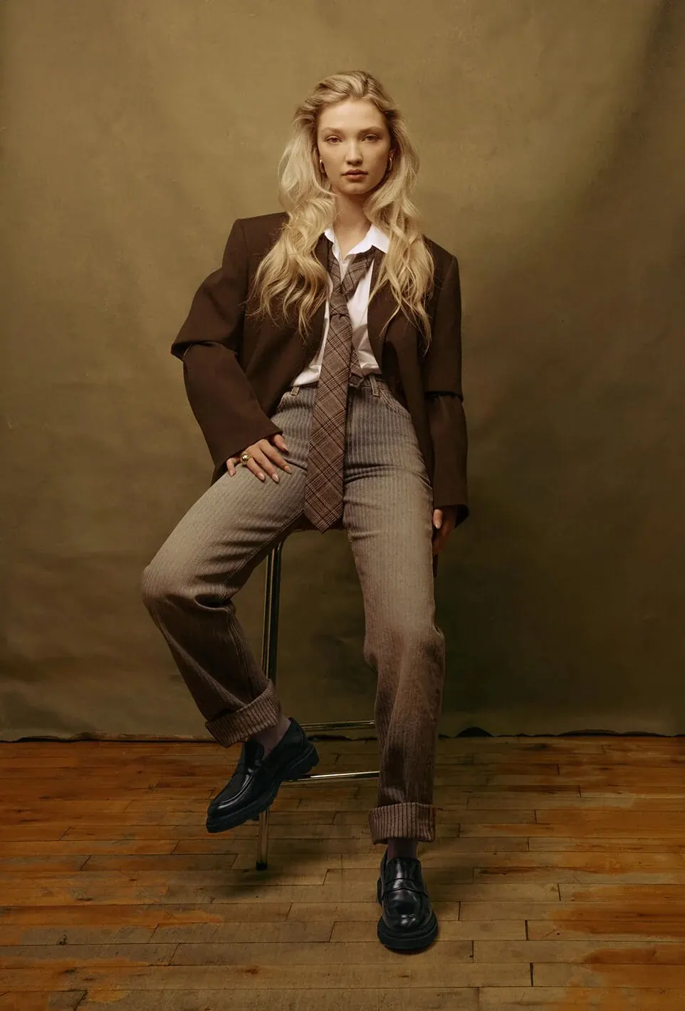 Woman in a brown blazer, tie and pinstripe trousers sitting on a stool in front of an olive-coloured photo backdrop, wooden floor
