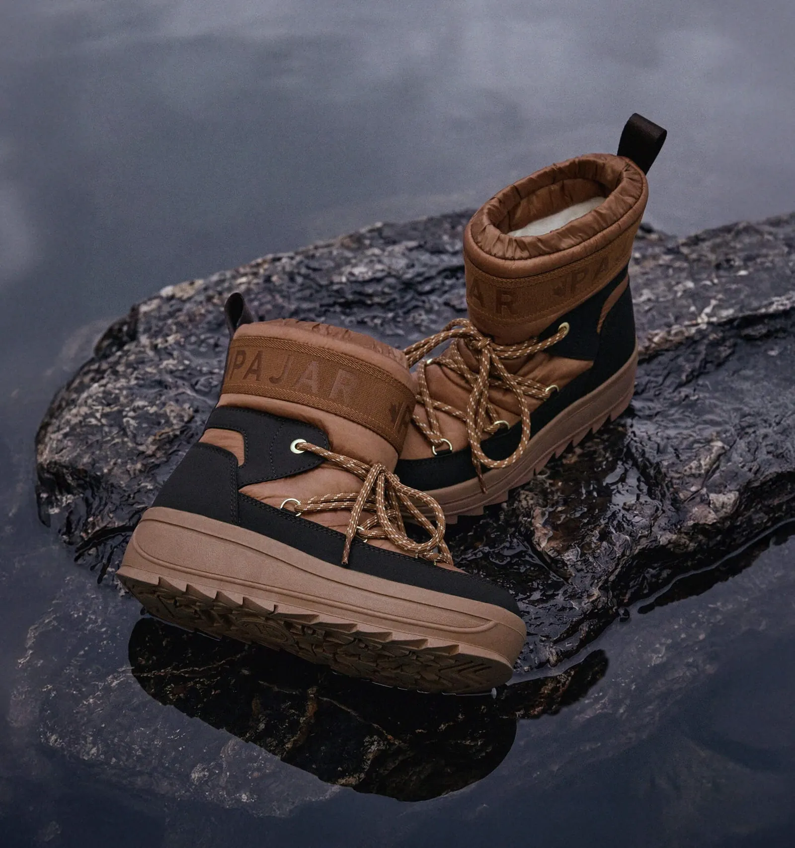 A pair of brown lace-up winter boots with a quilted shaft and Pajar lettering on a wet rock in the water