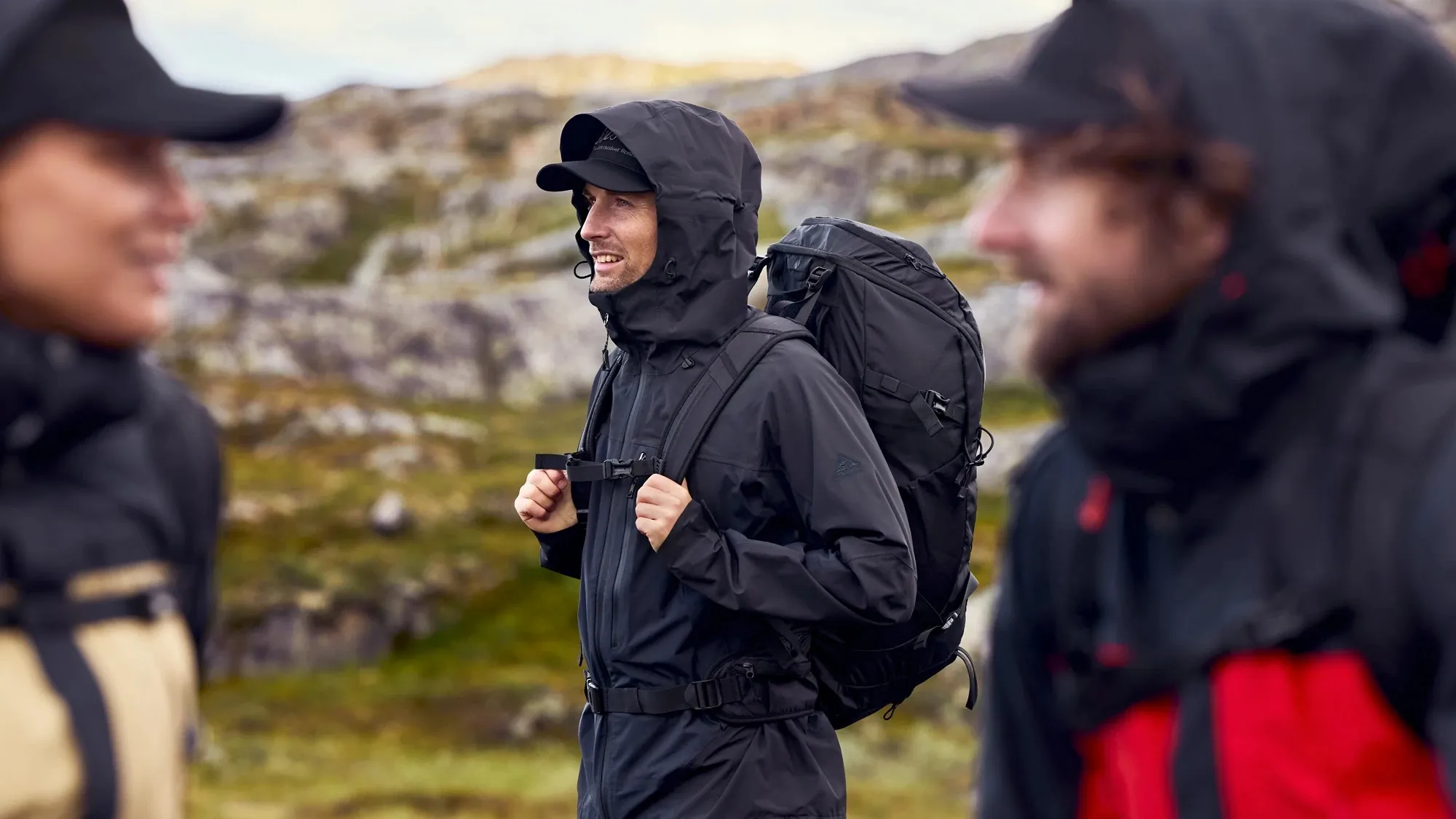 Lachender Wanderer mit Kapuze, schwarzer Shelljacke und Rucksack im Fjell, vorn unscharf zwei weitere Wanderer