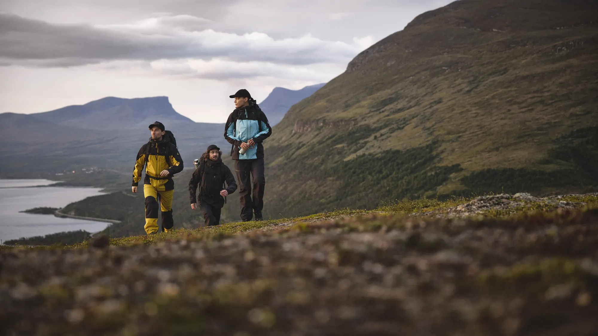Drei Wanderer in Outdoorjacken auf einem Höhenrücken über einem See, dahinter das Bergtal Lapporten in Schwedisch-Lappland