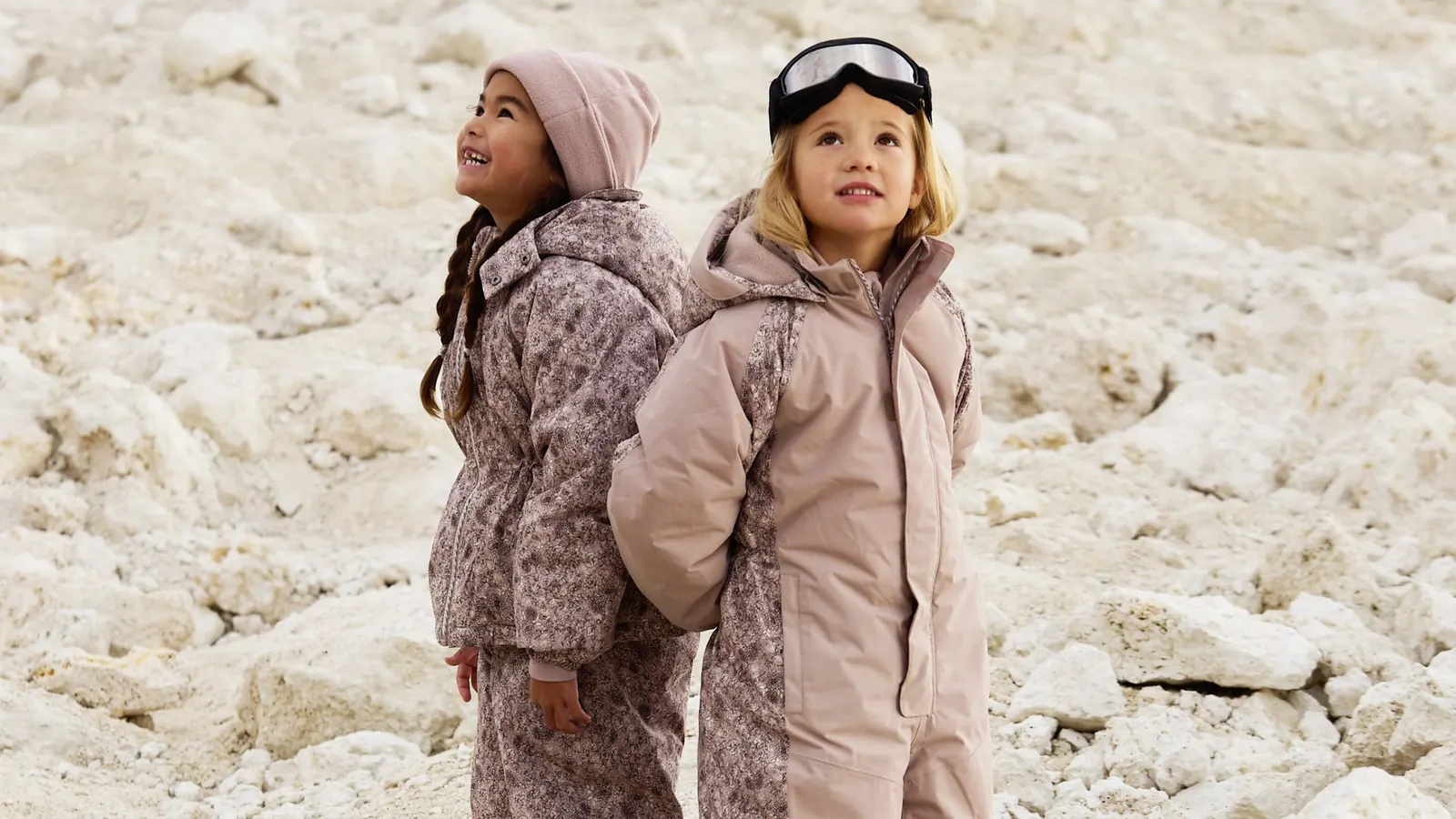 Zwei Mädchen in rosafarbener Winterkleidung stehen auf hellen Felsen, eines trägt eine Skibrille auf der Mütze