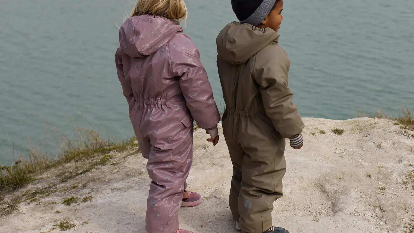 Zwei Kinder in einteiligen Schneeanzügen, eines in Rosa, eines in Oliv, stehen von hinten gesehen auf einer Felskante am Wasser