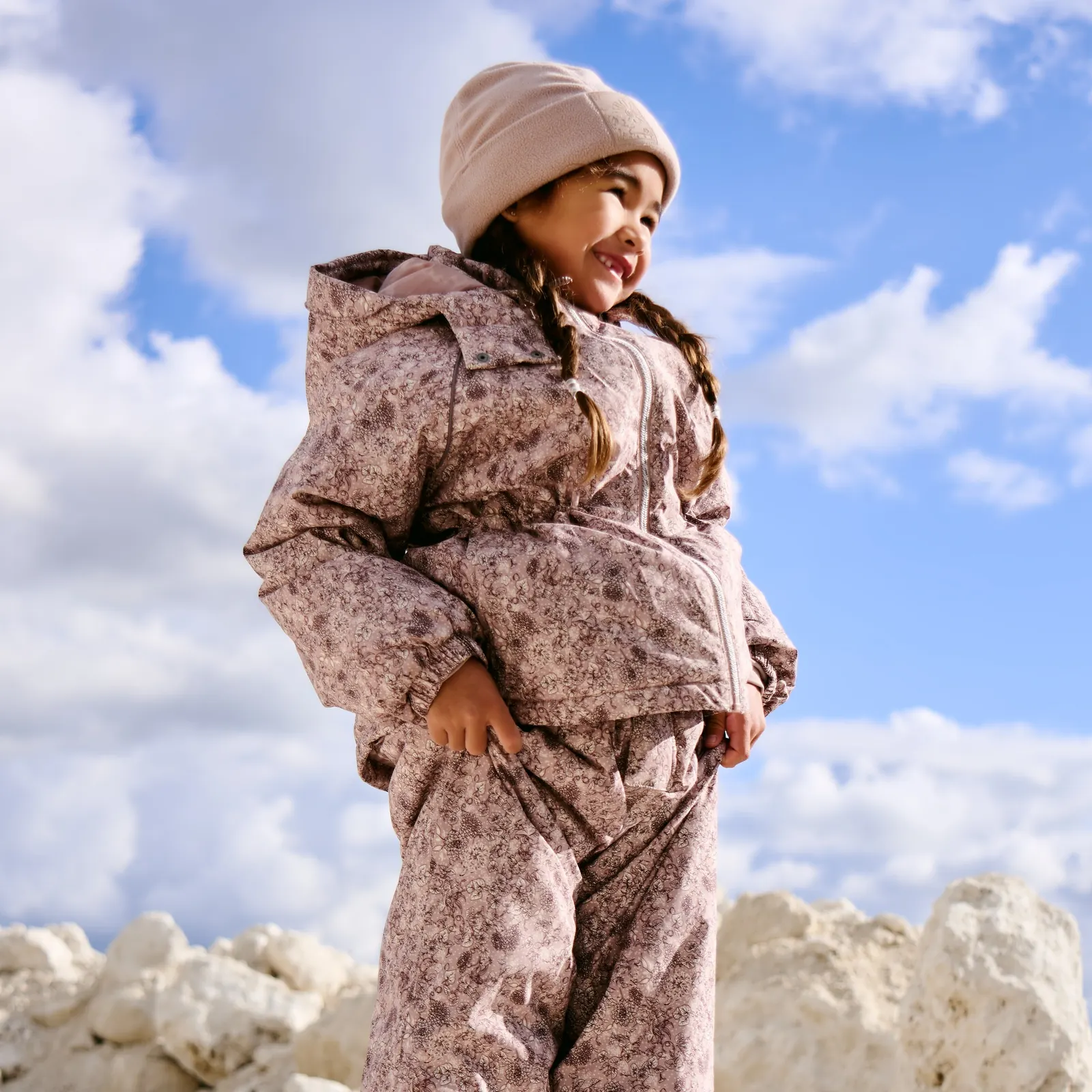 Mädchen mit Mütze in Skijacke und Skihose mit kleinem Blumenmuster vor blauem Himmel