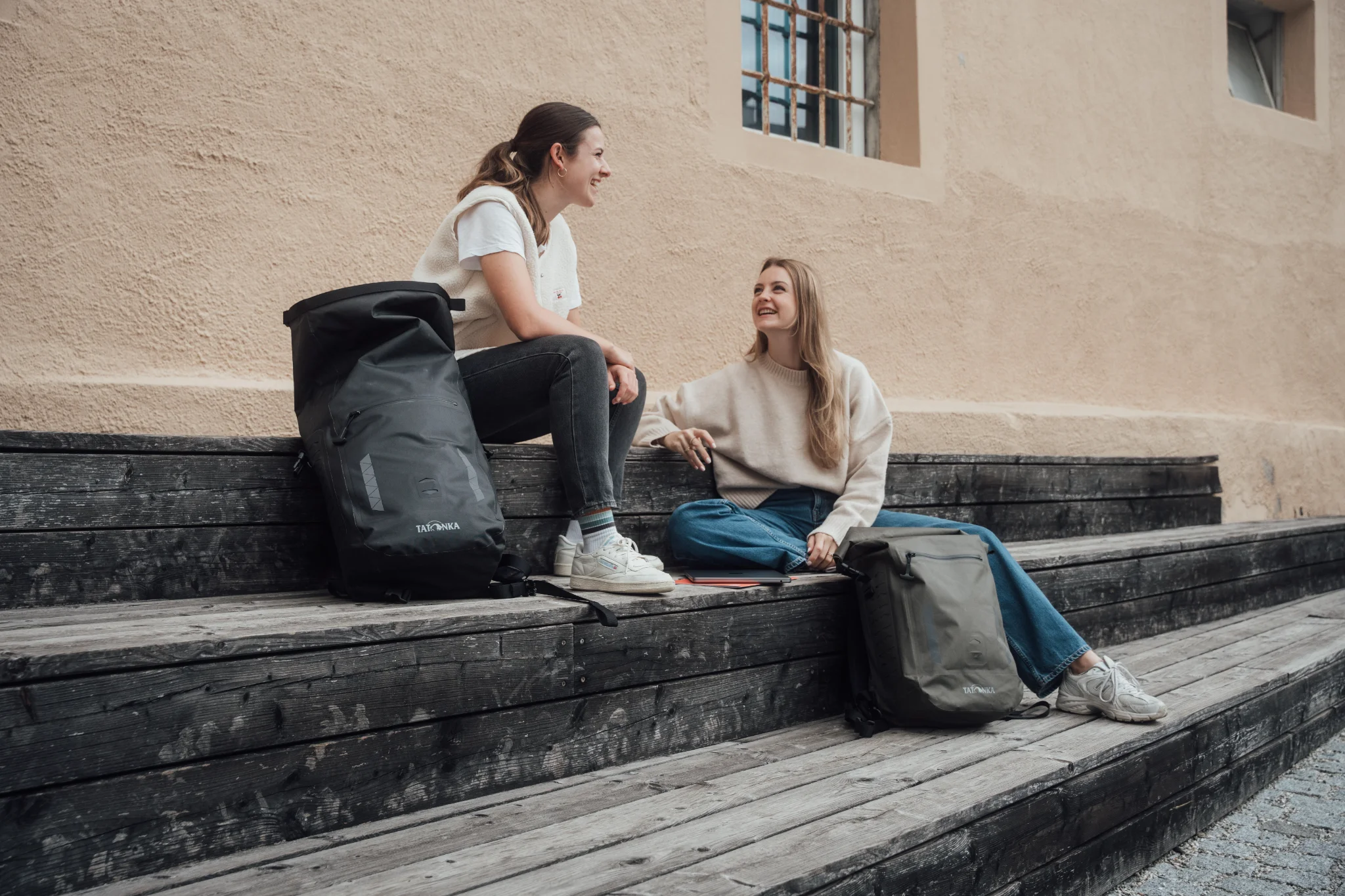 Two young women sitting on wooden steps with a black and an olive Tatonka roll-top rucksack