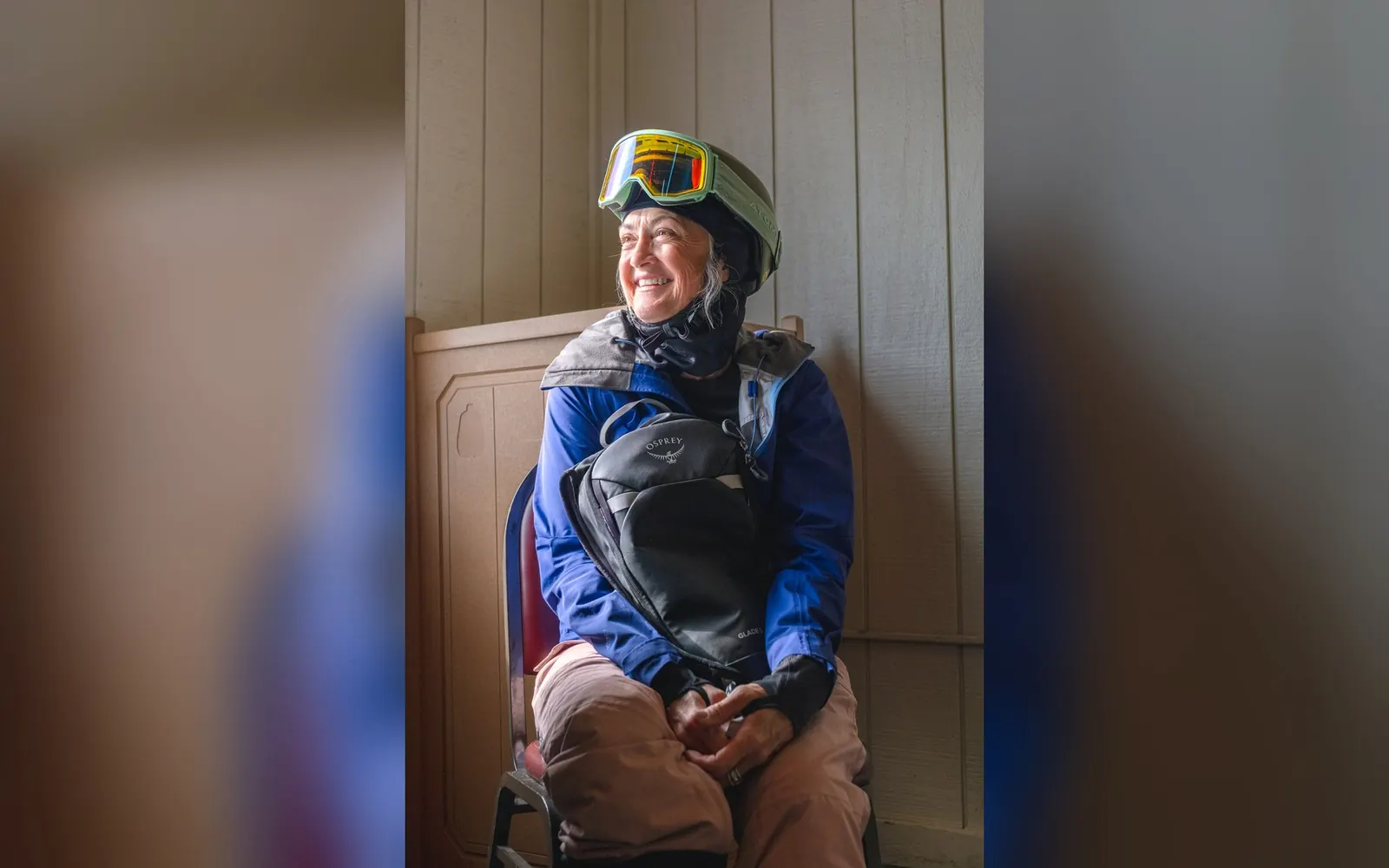 Lächelnde Skifahrerin mit Helm und Skibrille, auf dem Schoß ein schwarzer Osprey Glade 20