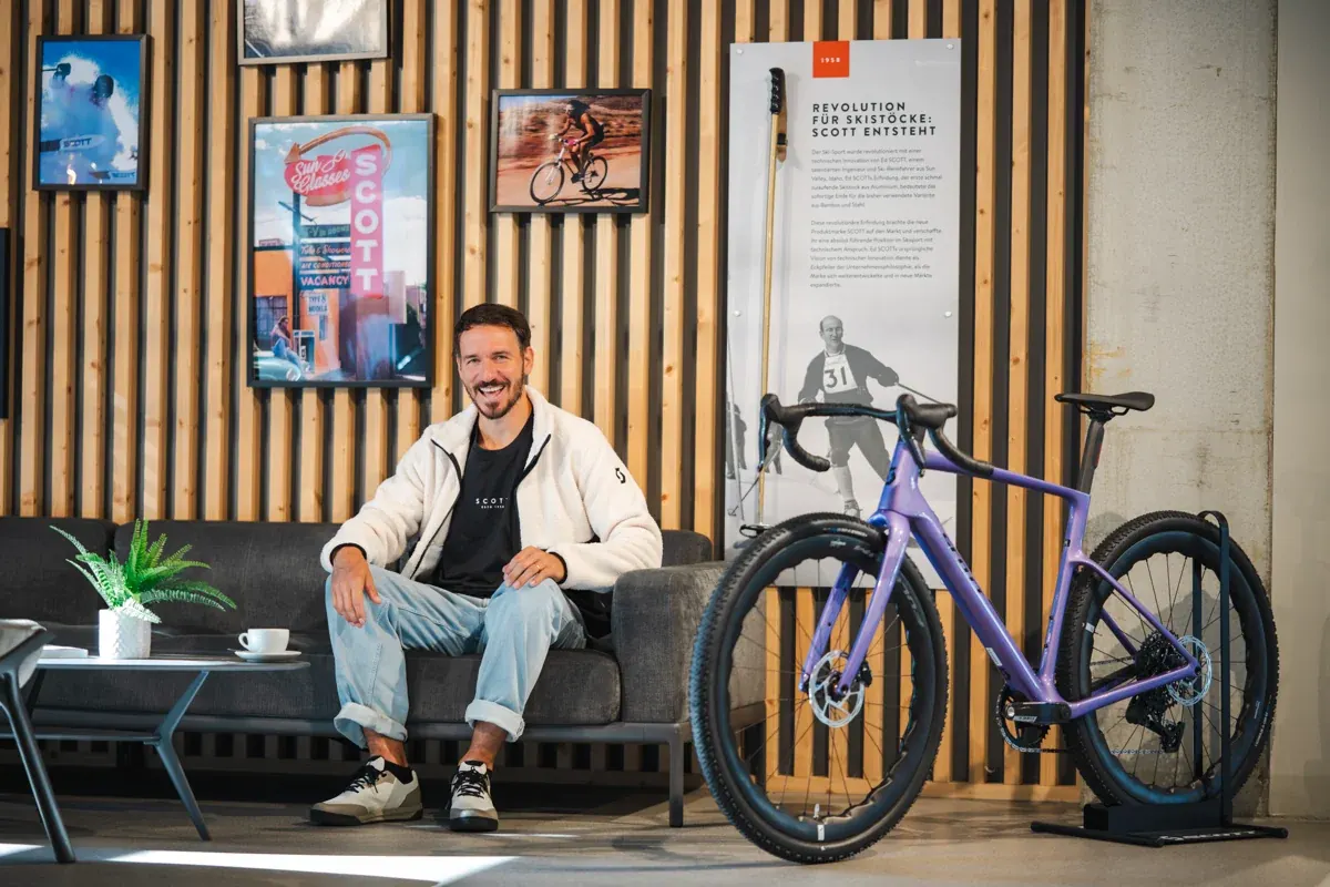 Felix Neureuther sitzt in einem Scott-Laden auf einem Sofa neben einem Rennrad, dahinter ein Plakat zur Geschichte der Scott-Skistöcke