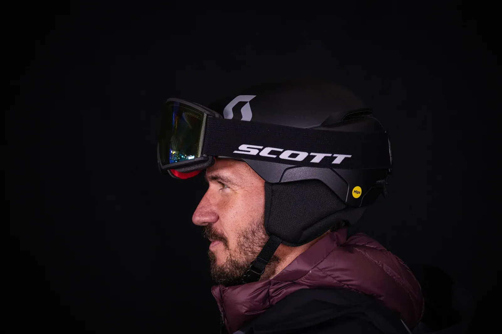 Felix Neureuther im Profil mit schwarzem Scott-Helm, Skibrille auf dem Helm und weinroter Jacke vor dunklem Hintergrund