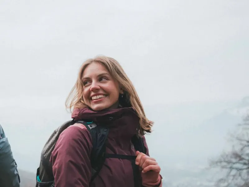 Lachende Wanderin in Schöffel-Jacke mit Rucksack vor nebligen Bergen