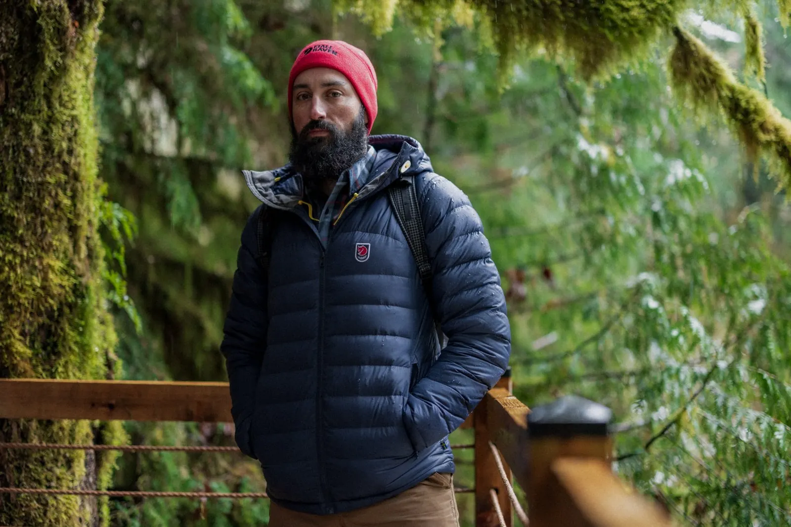 Man with a red beanie in a dark blue Fjällräven down jacket on a wooden boardwalk in the forest