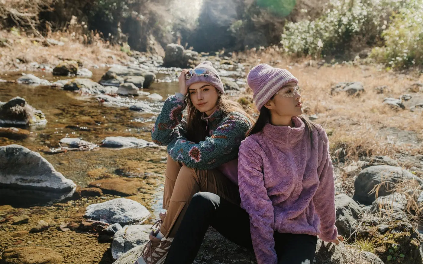 Two women in fleece jackets and beanies sitting by a stream