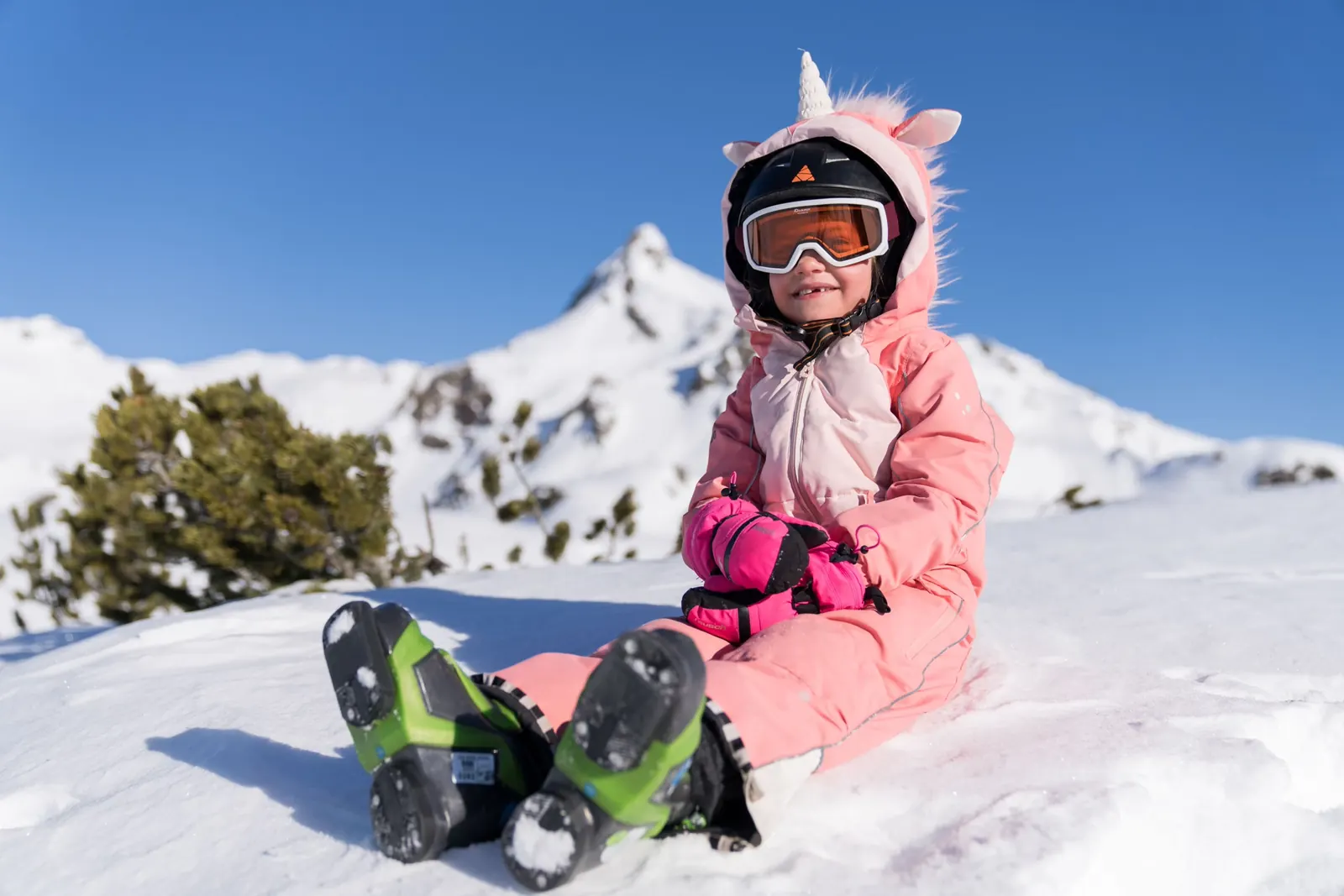 Kind mit Skihelm und Skibrille sitzt lachend im Schnee vor Bergpanorama