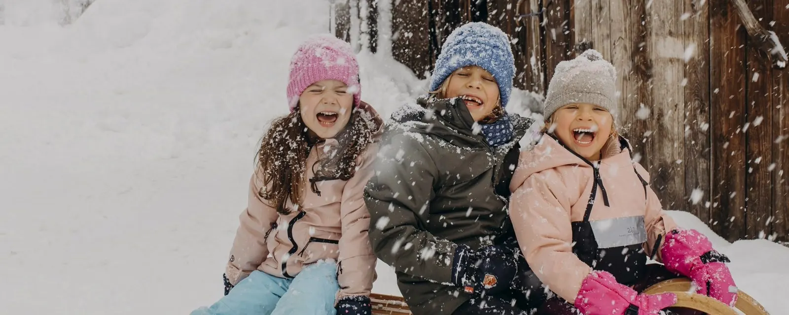 Drei Kinder lachen beim Schlittenfahren im Schnee vor einem Holzhaus