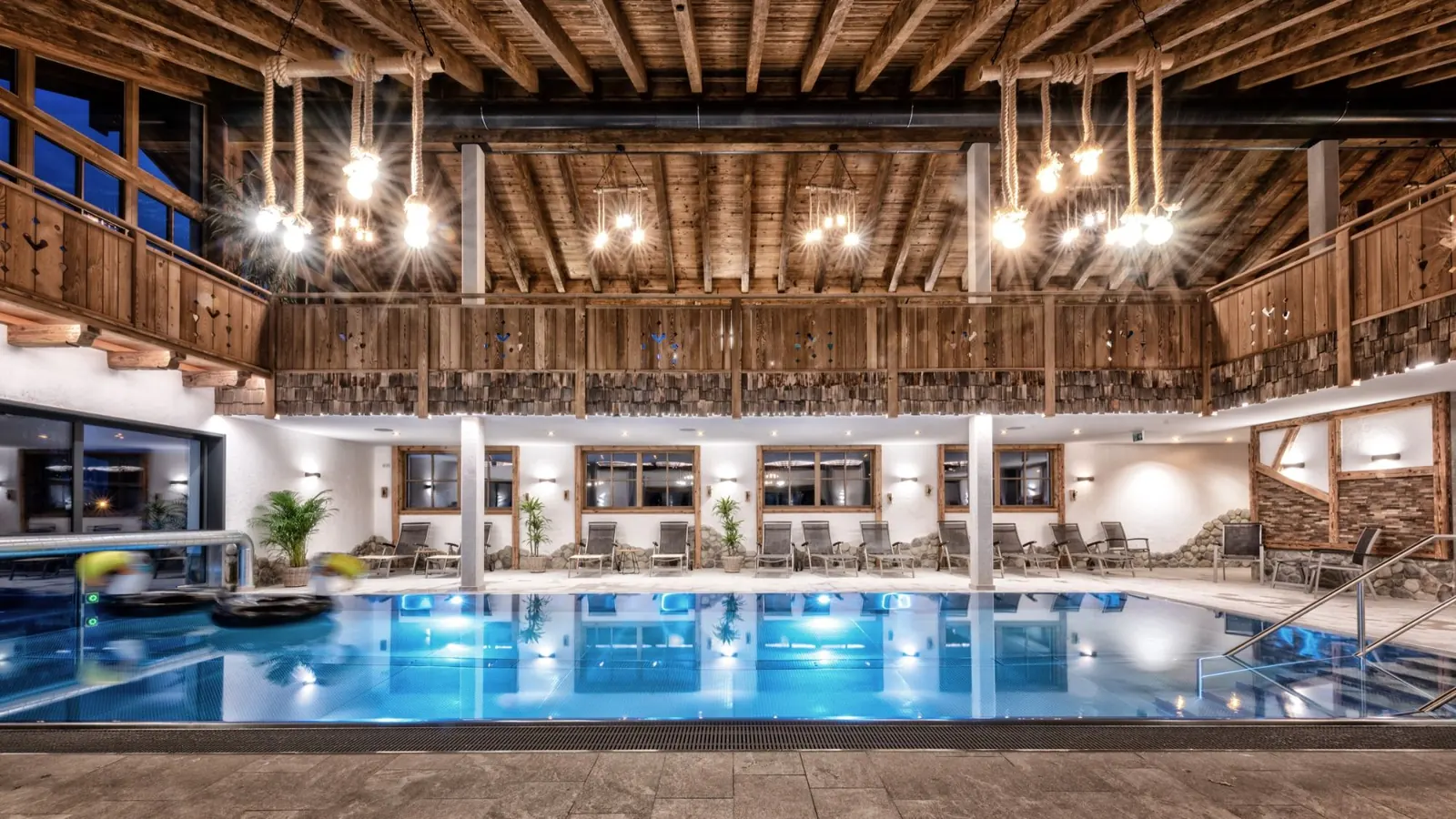 Beleuchteter Innenpool mit Holzbalkendecke und Liegestühlen im Mühlpointhof