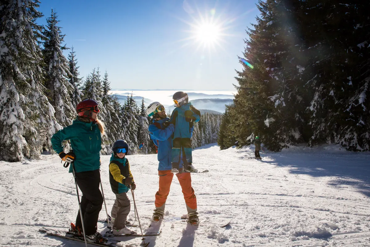 Eine Familie mit zwei Kindern steht in Skiausrüstung auf einer Piste zwischen verschneiten Fichten