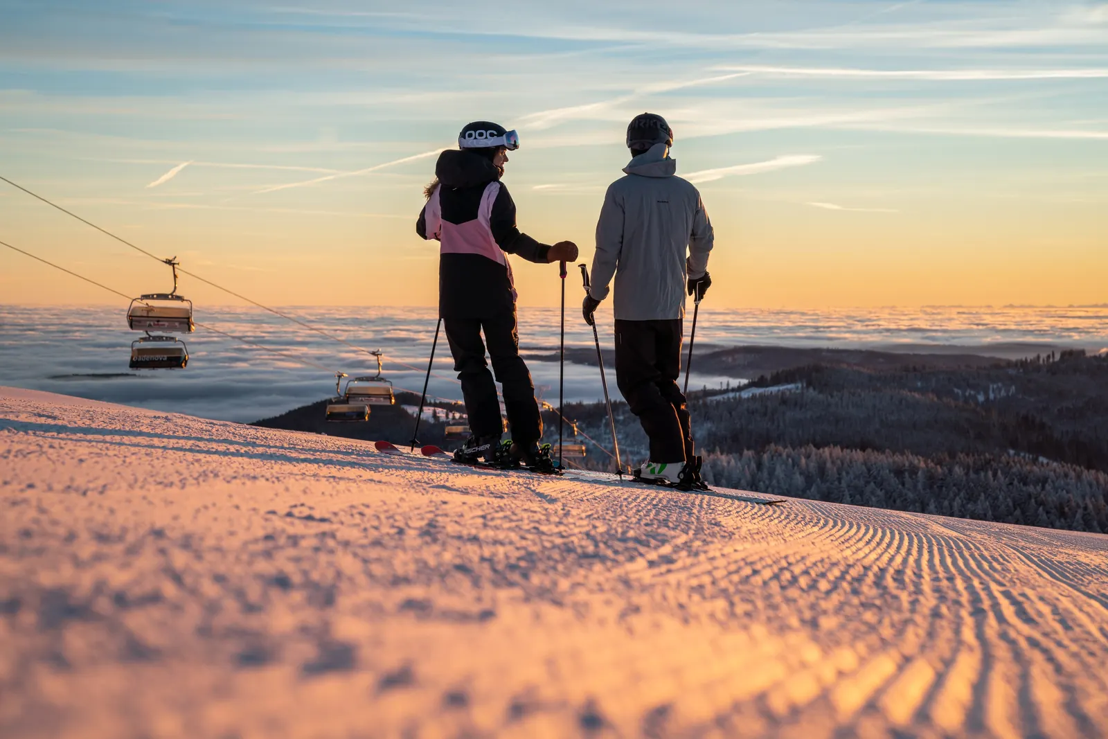 Zwei Skifahrer stehen am Abend auf einer präparierten Piste am Feldberg, dahinter Sessellift und Nebelmeer über dem Schwarzwald