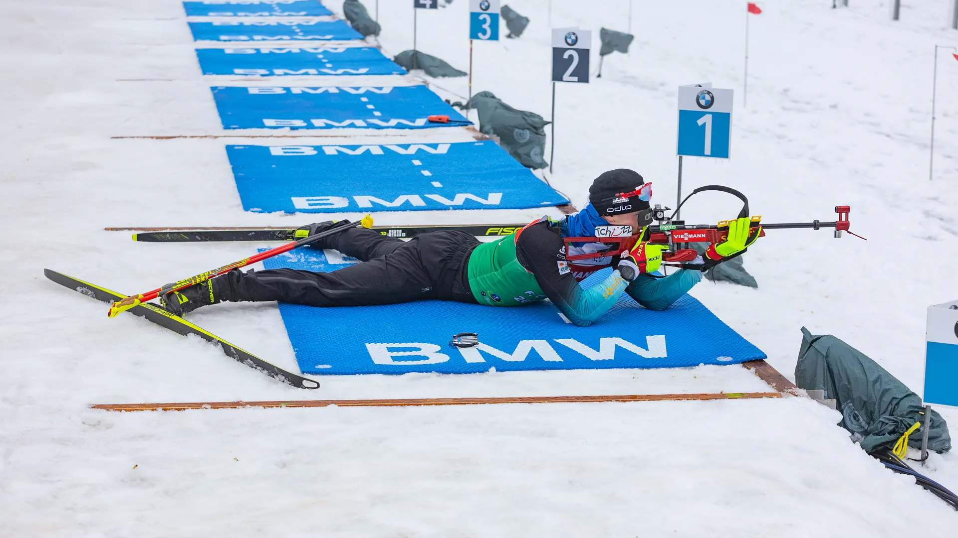 Biathlet liegt auf der Schießmatte am Schießstand und zielt mit dem Gewehr auf die Scheiben, im Hintergrund die Startnummernschilder und Matten der Nachbarstände
