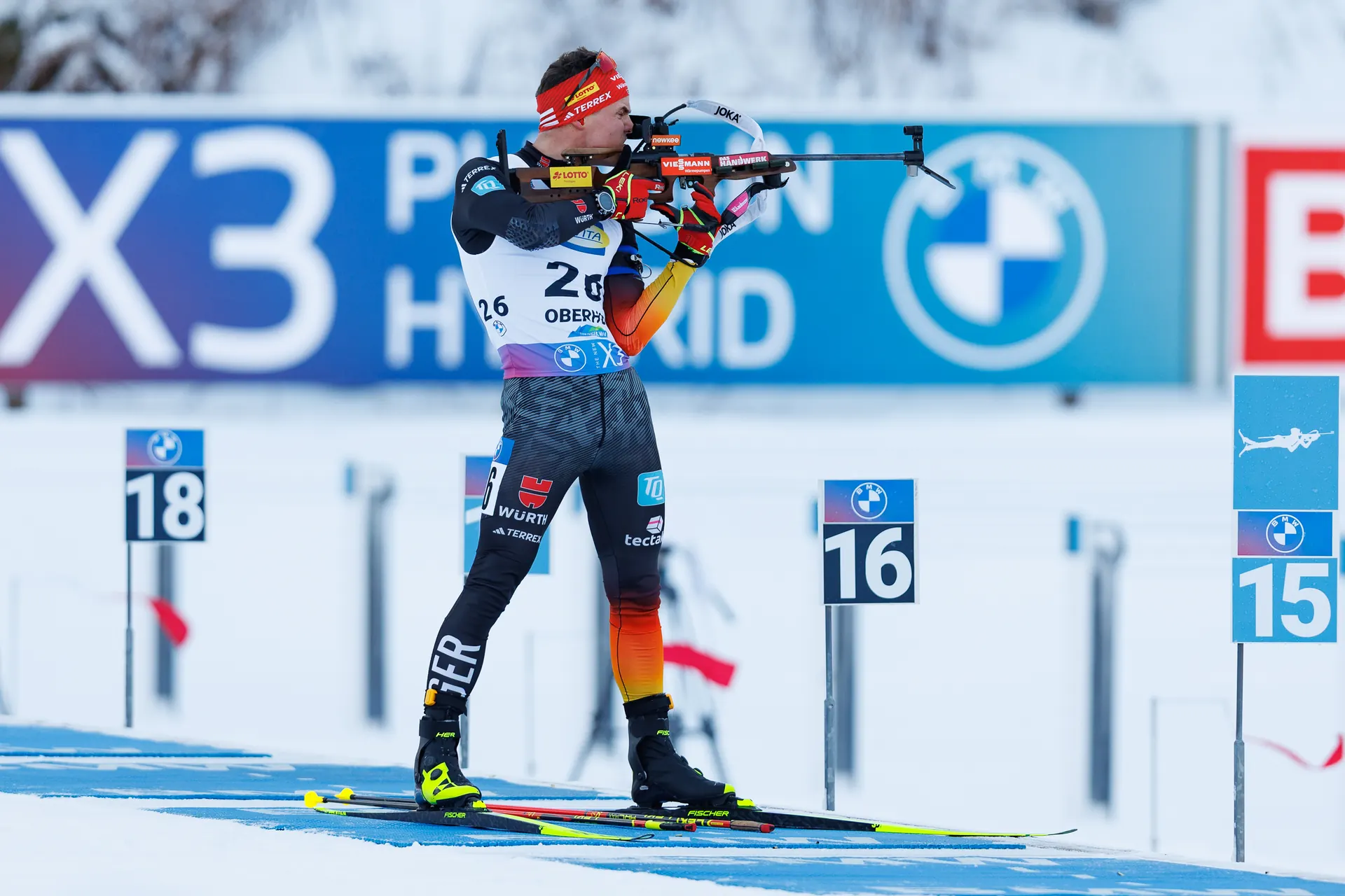 Biathlet liegt auf der Schießmatte am Schießstand und zielt mit dem Gewehr auf die Scheiben, im Hintergrund die Startnummernschilder und Matten der Nachbarstände