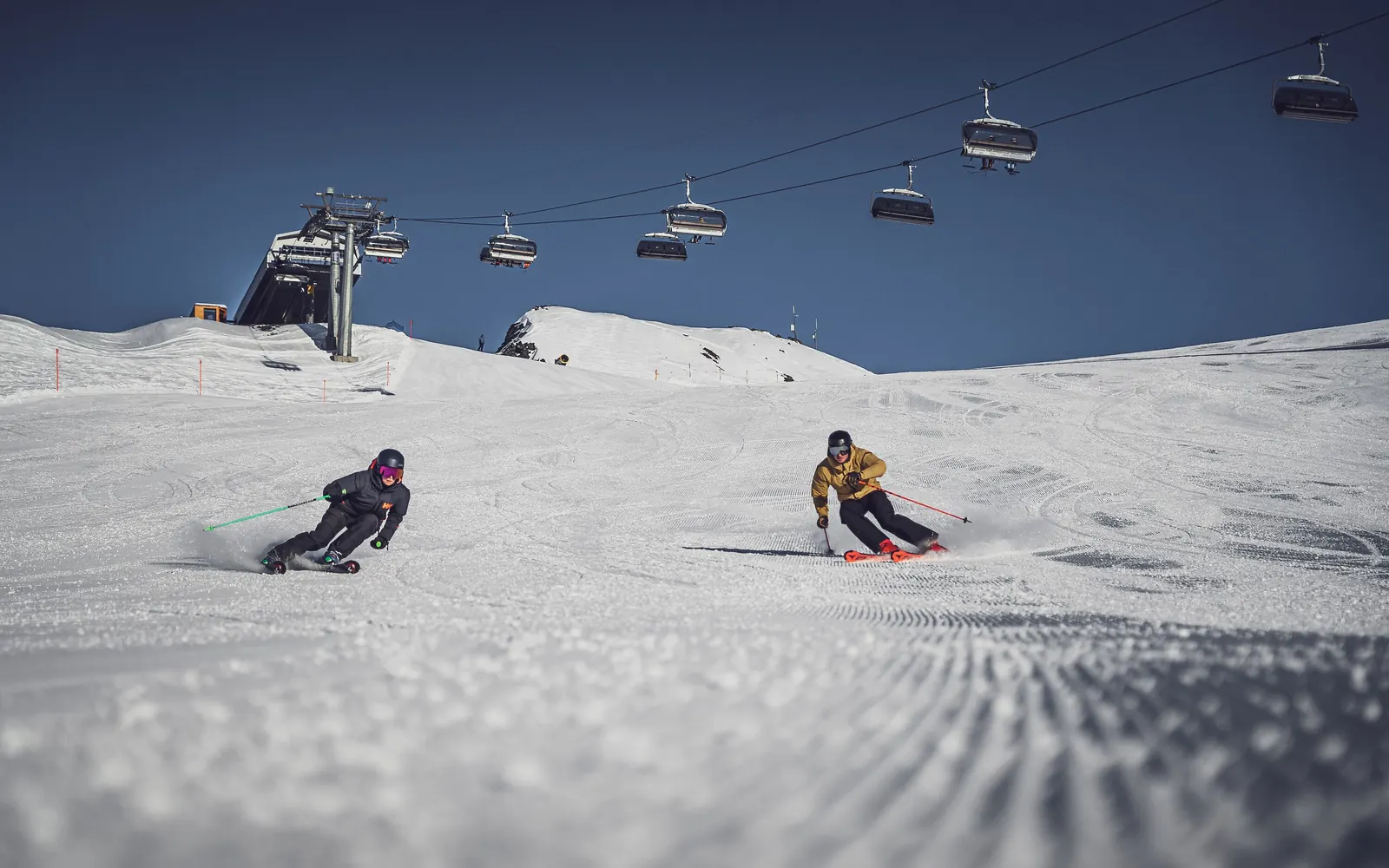 Zwei Skifahrende carven auf einer frisch präparierten Piste, über ihnen eine Sesselbahn