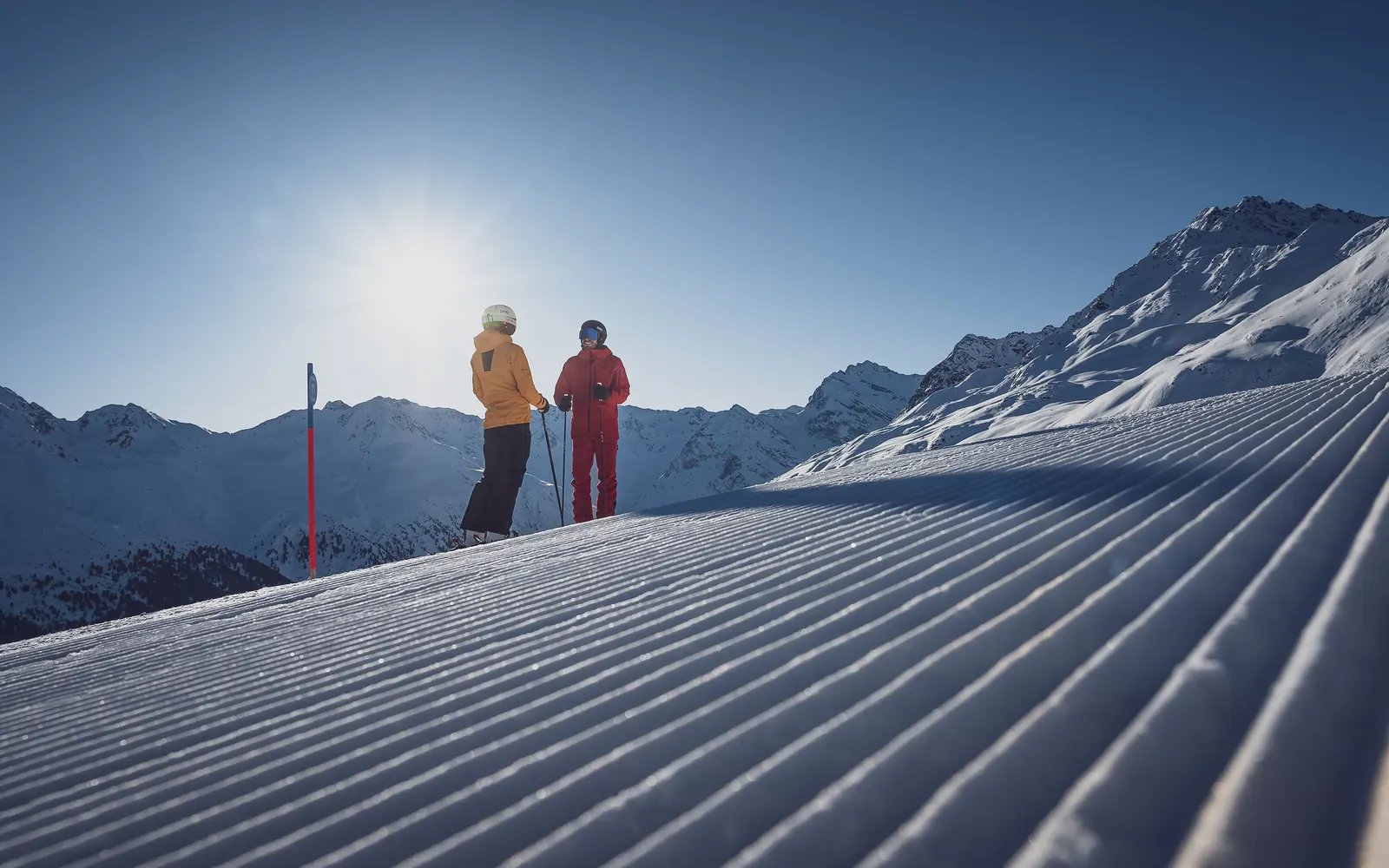 Zwei Skifahrende stehen im Gegenlicht am Rand einer frisch präparierten Piste, dahinter verschneite Gipfel