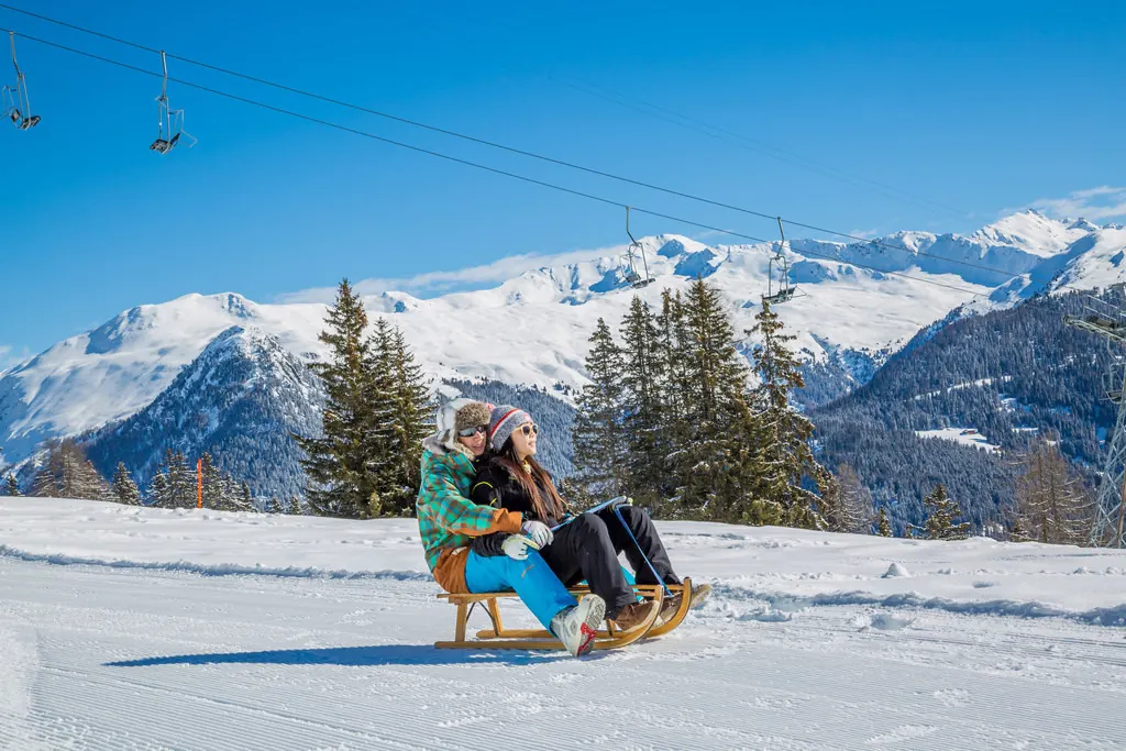 Zwei Personen fahren auf einem Holzschlitten über eine präparierte Schneebahn, im Hintergrund Sessellift und verschneite Berge