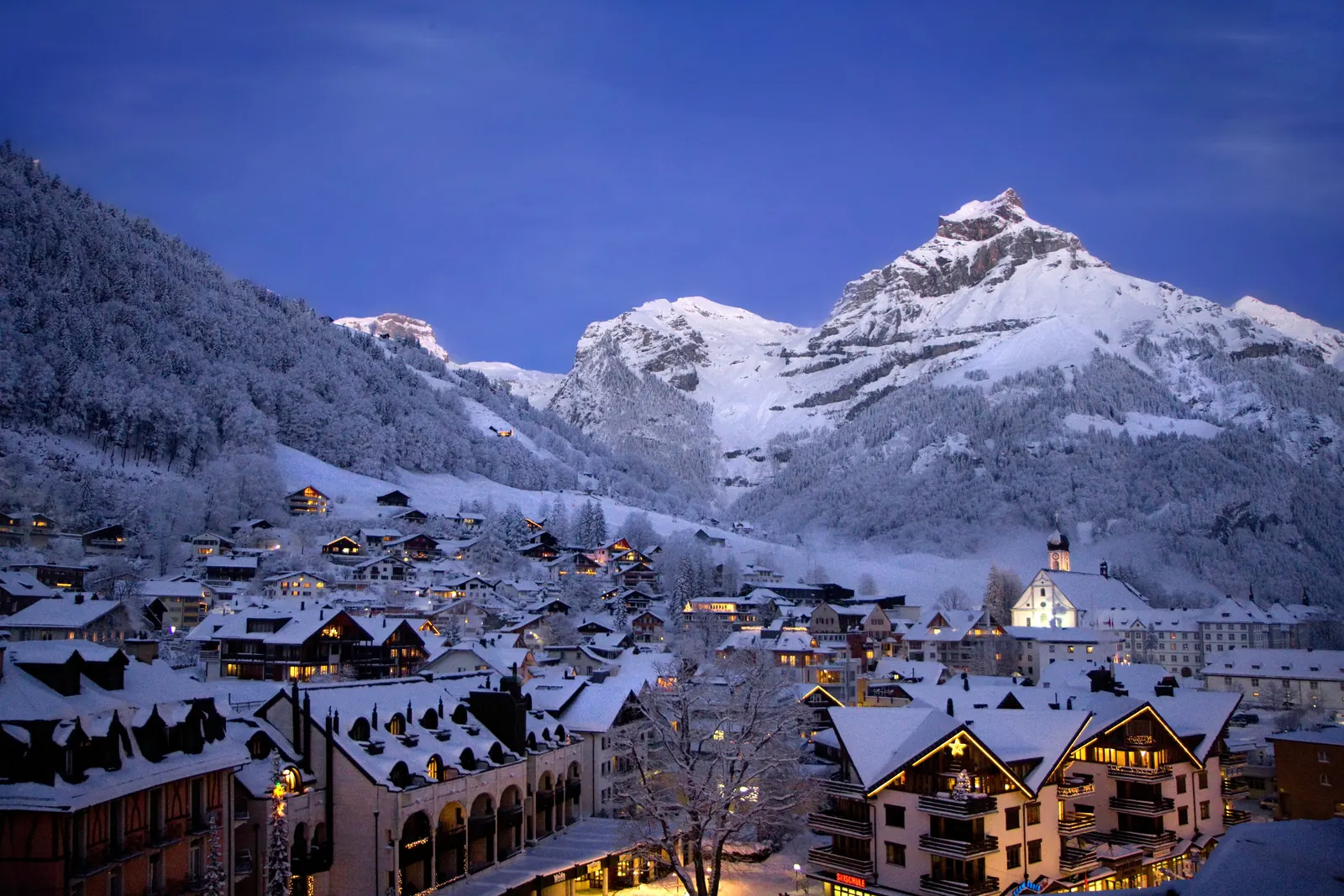 Verschneites Dorf Engelberg in der Abenddämmerung mit beleuchteten Häusern, dahinter schneebedeckte Felsgipfel