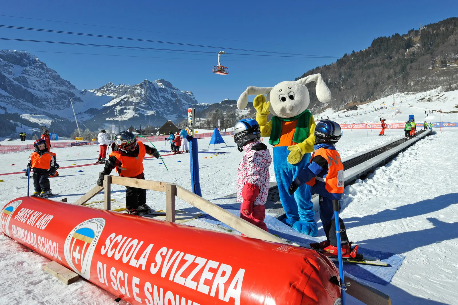 Kinder in Skikleidung mit Helm stehen an einem Förderband im Übungsgelände, daneben ein Skischul-Maskottchen