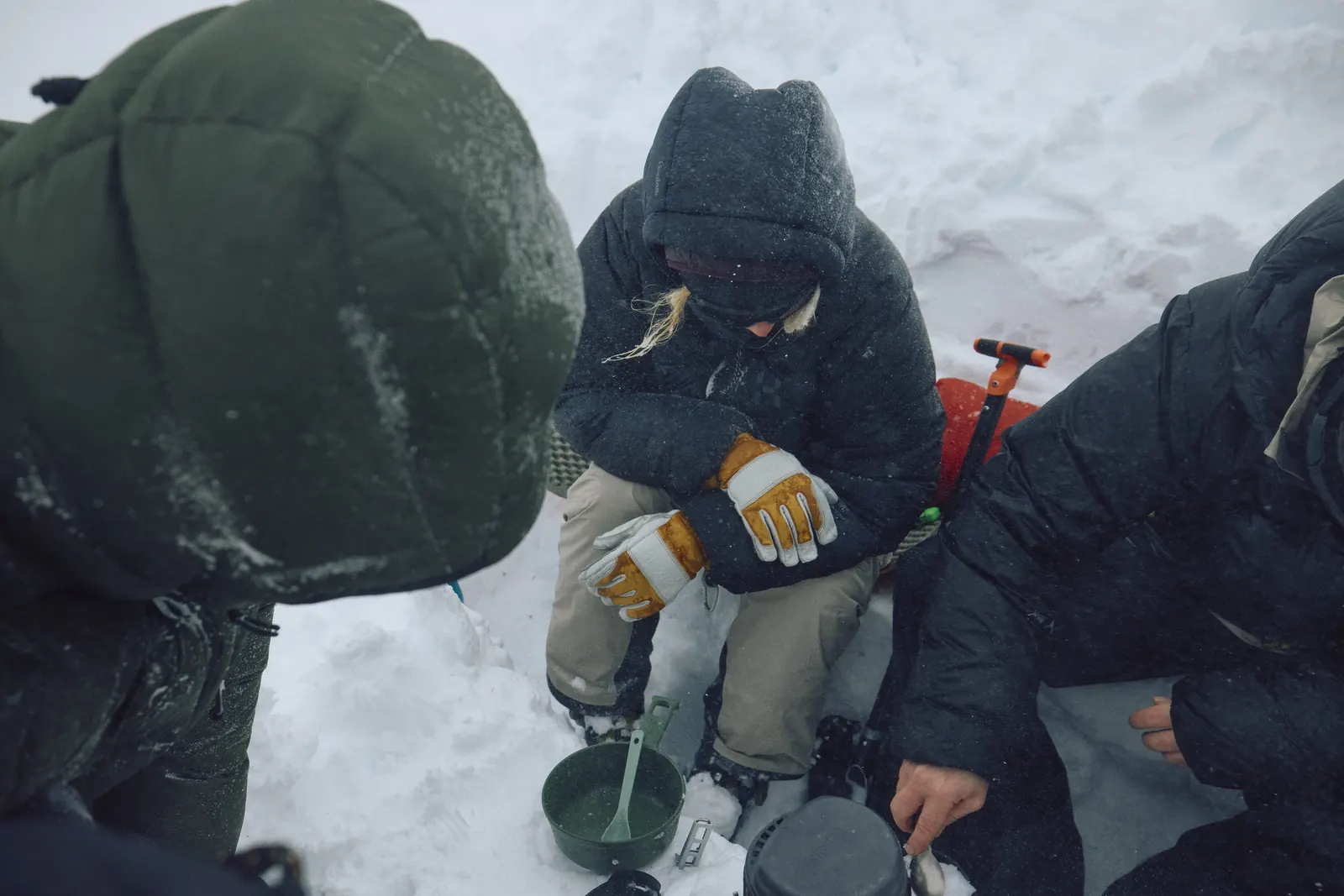 Three people in down jackets sitting in the snow around a stove
