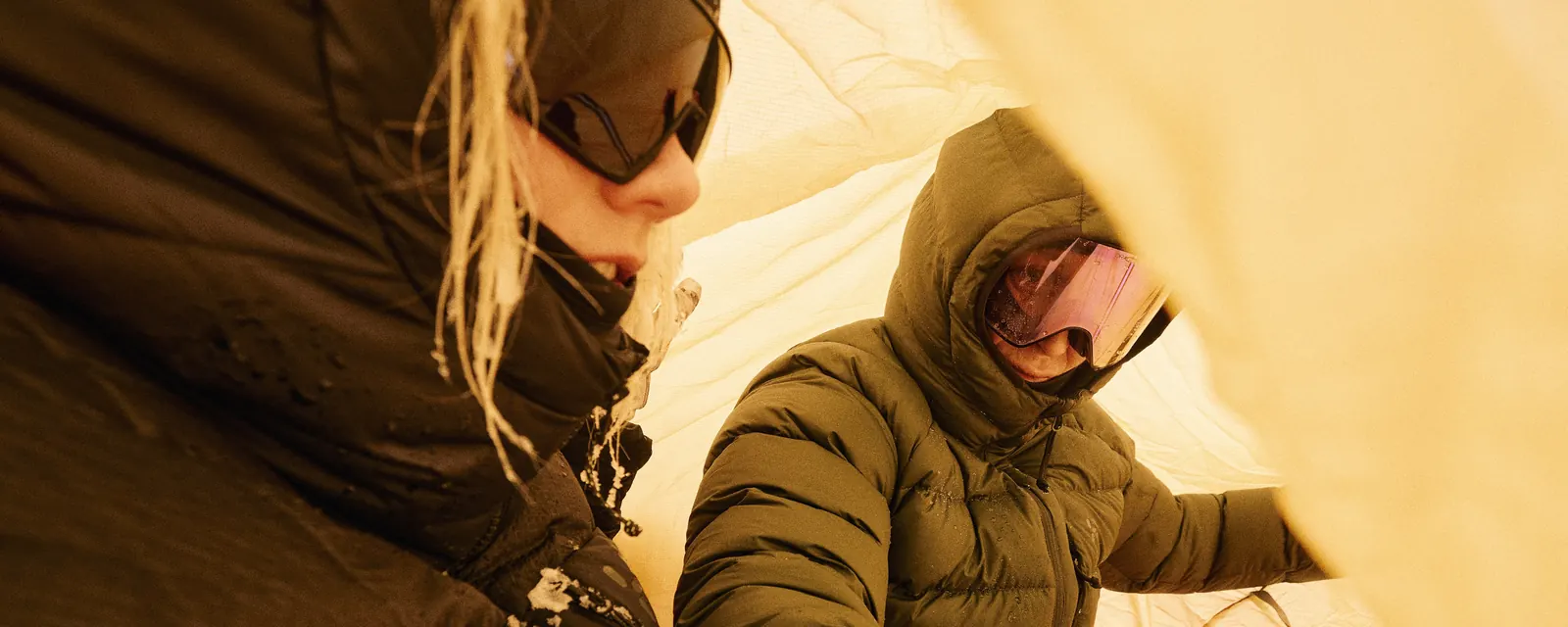 Zwei Personen in Haglöfs Daunenjacken mit Kapuze und Skibrille im gelben Zelt