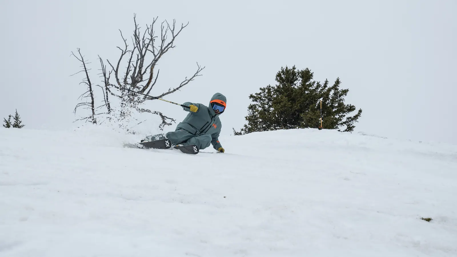 Skier in petrol-coloured Helly Hansen ski wear making a deep turn in the snow next to a bare bush