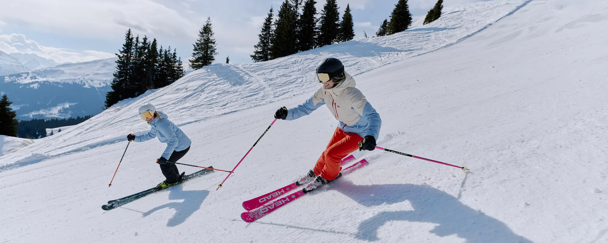 Zwei Skifahrerinnen in Helly-Hansen-Skijacken fahren nebeneinander eine Piste hinunter