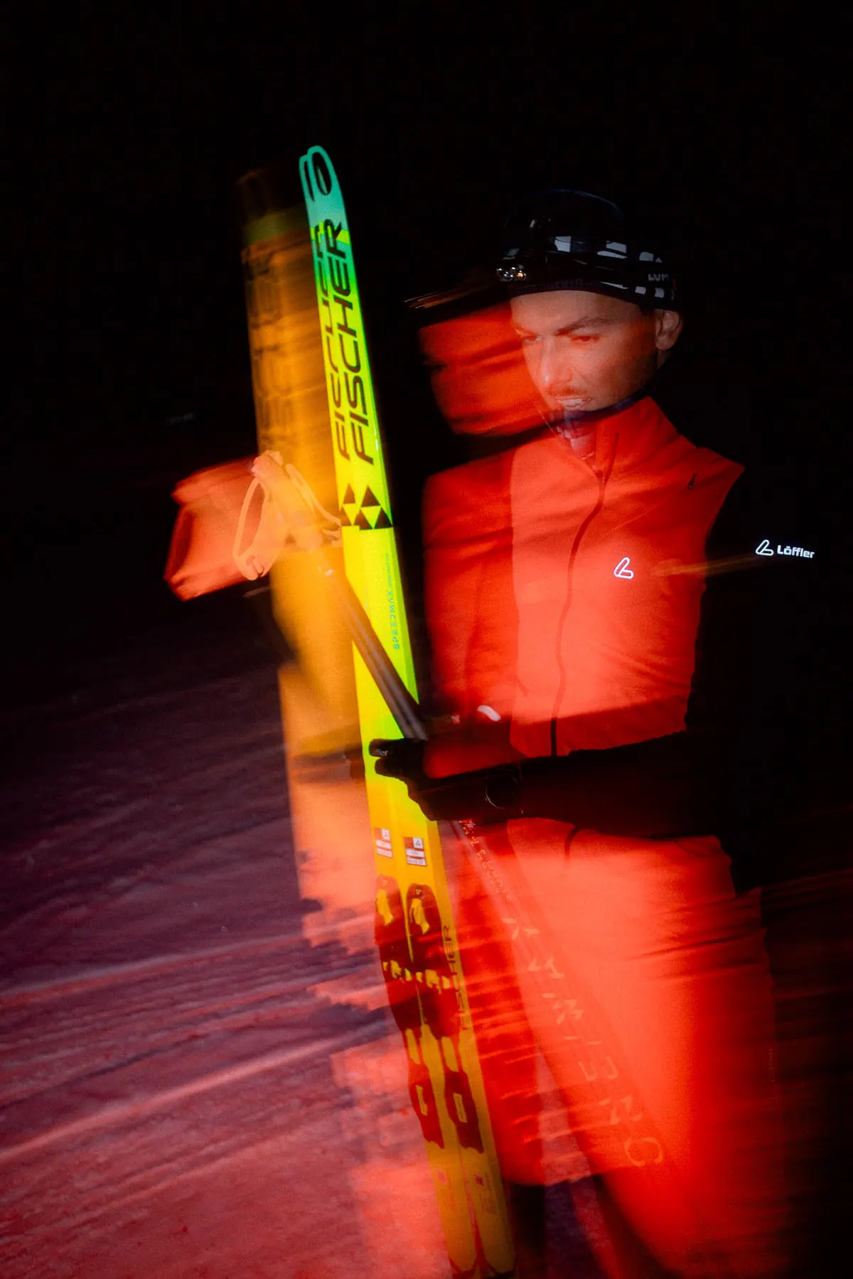 Langläufer in roter Jacke mit Löffler-Logo am Ärmel, Skier und Stöcke in der Hand, nachts mit Bewegungsunschärfe