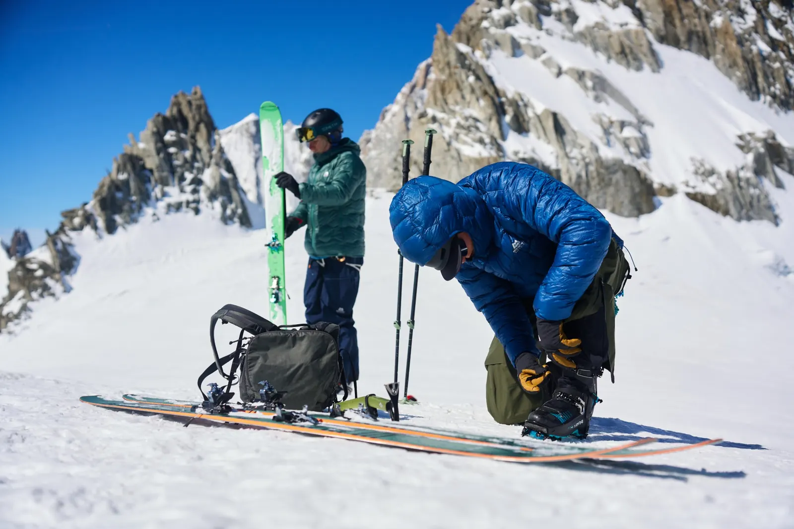 Ski tourer in a blue Xenith down jacket kneeling to adjust ski boots, a second person with skis in the background