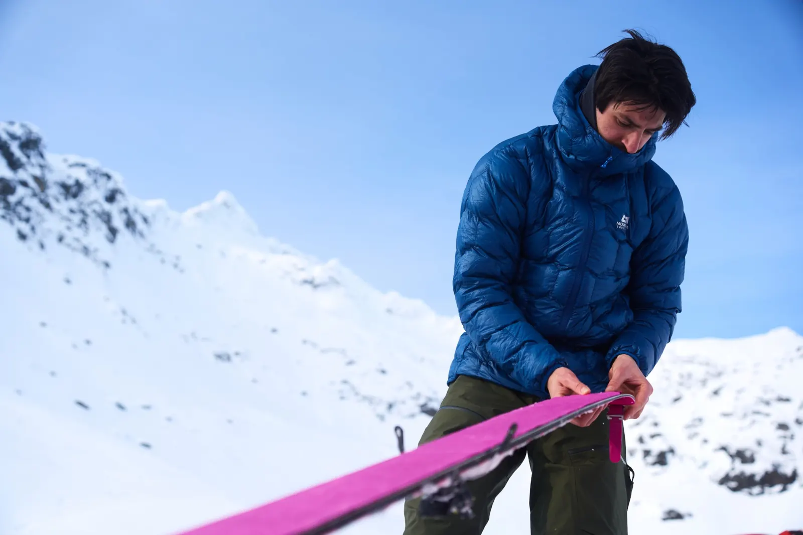 Ski tourer in a blue Xenith down jacket pulling the skins off his skis in front of a snowy mountain