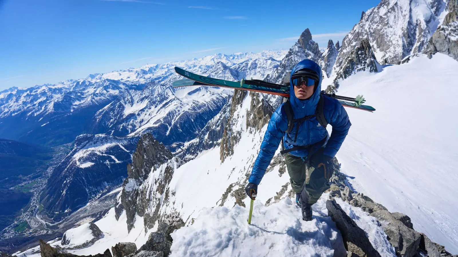 Mountaineer in a blue Mountain Equipment Xenith down jacket with skis on his pack climbing a snowy rocky ridge