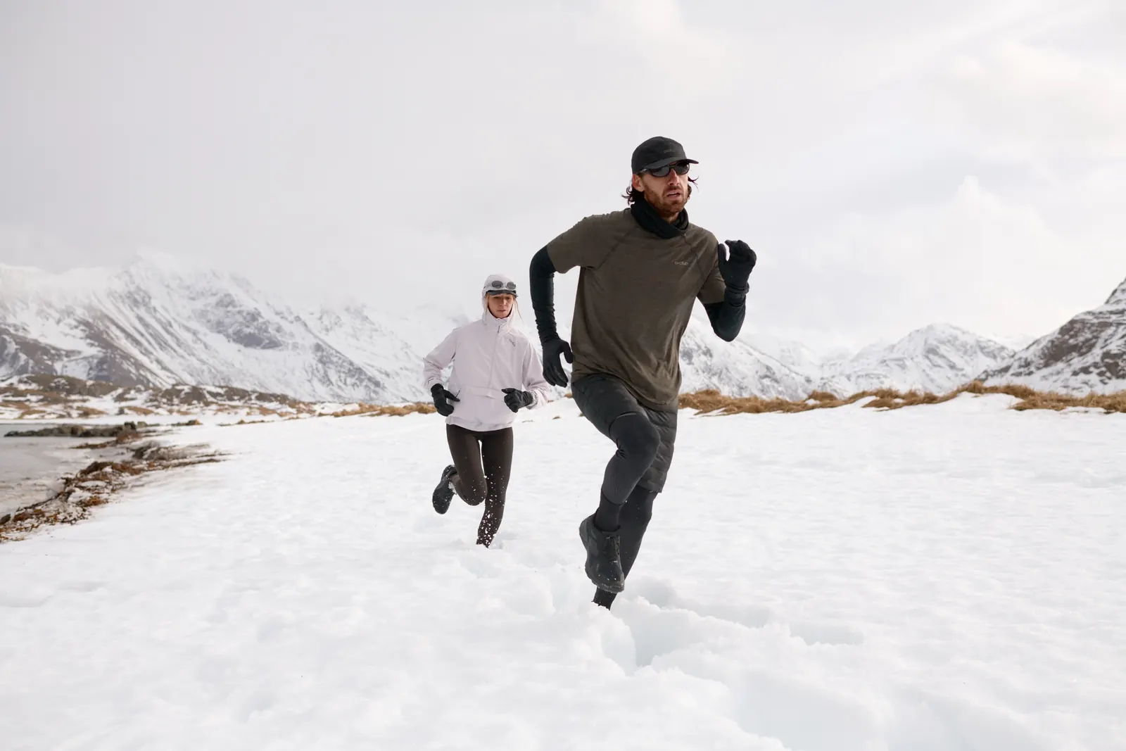Läufer und Läuferin laufen durch Schnee vor verschneiter Berglandschaft