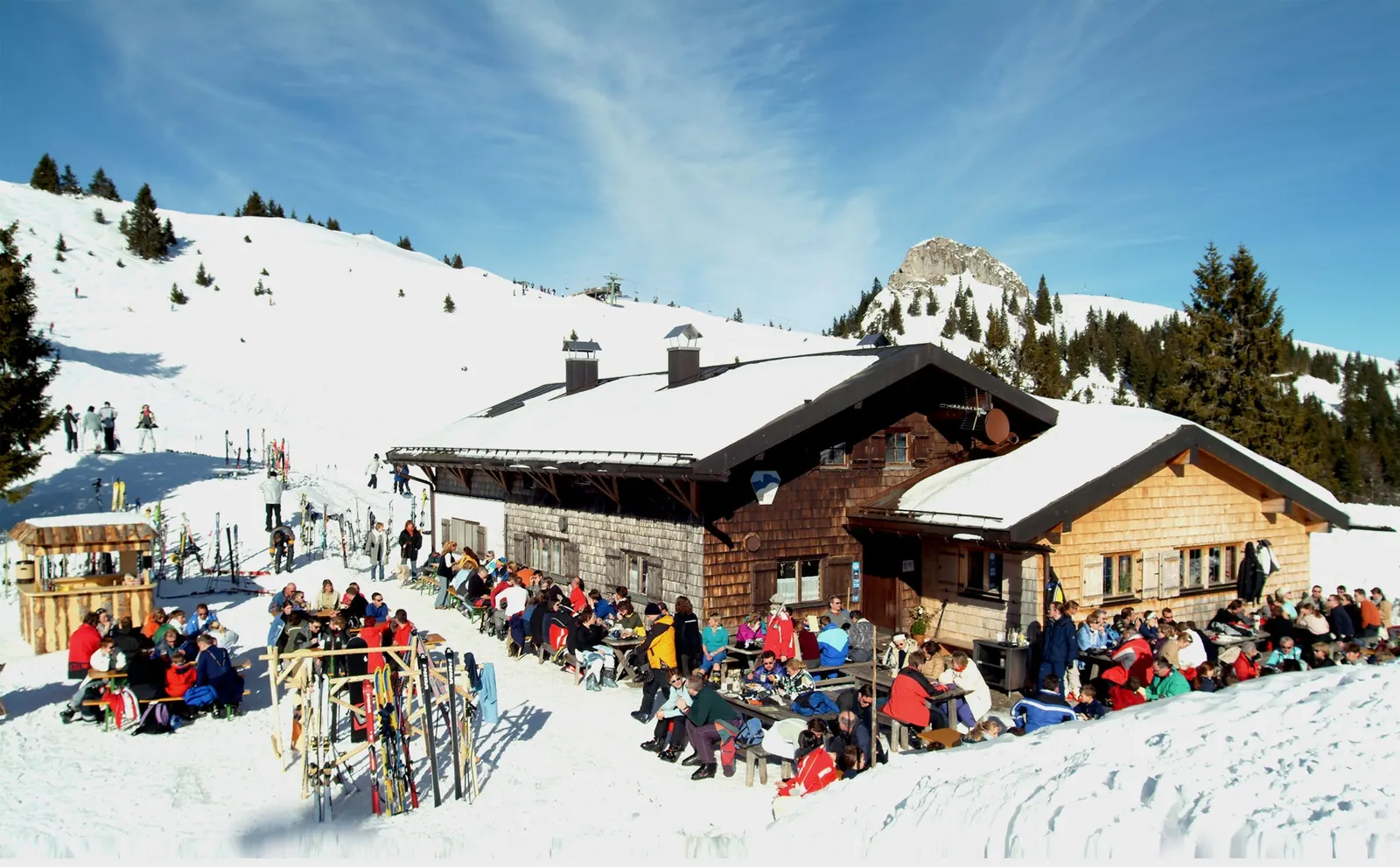 Voll besetzte Sonnenterrasse einer Holzhütte im Skigebiet Brauneck, davor abgestellte Ski