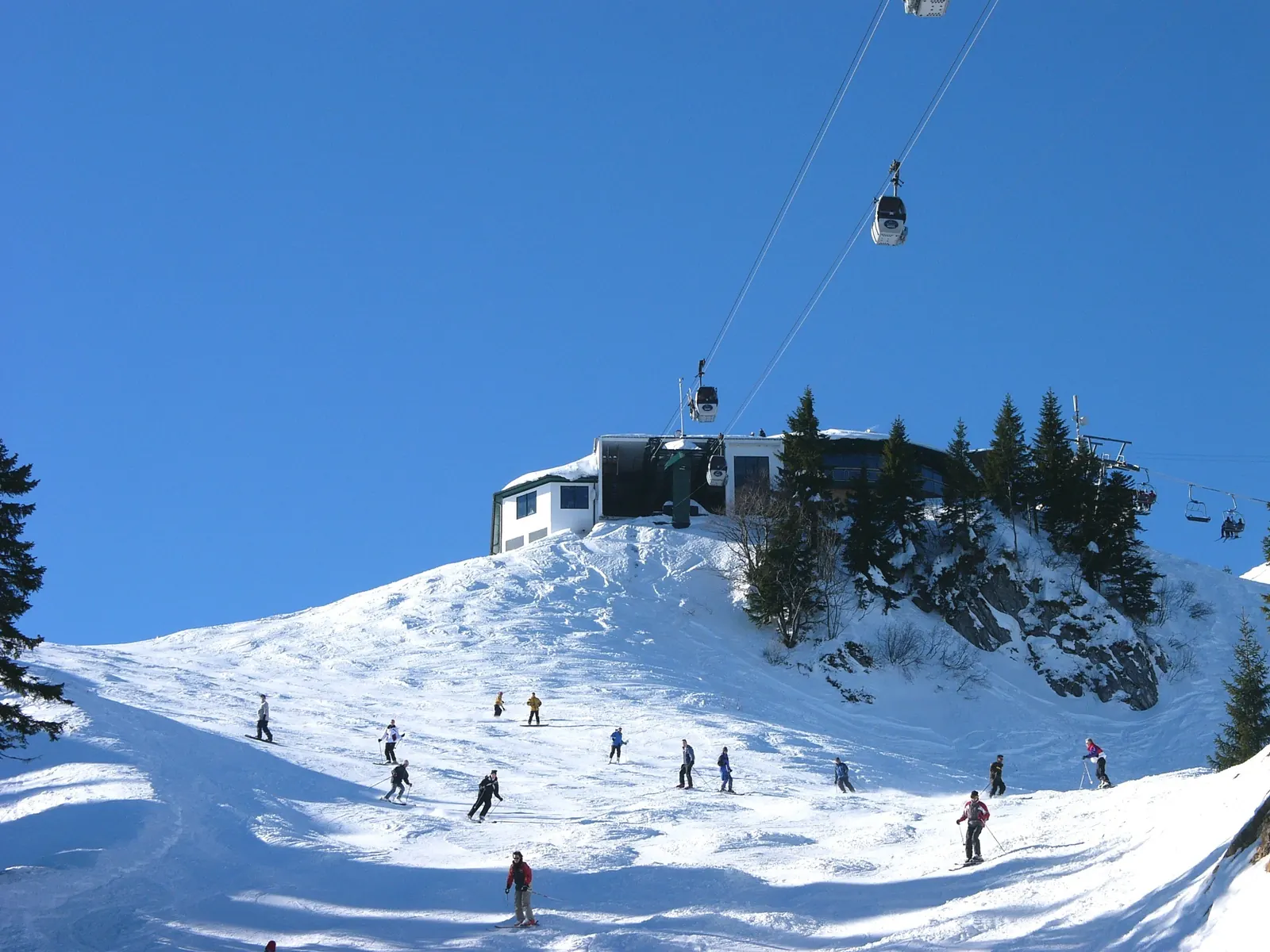 Skifahrer auf einer breiten Piste unterhalb der Bergstation am Brauneck, darüber eine Gondel der Kabinenbahn