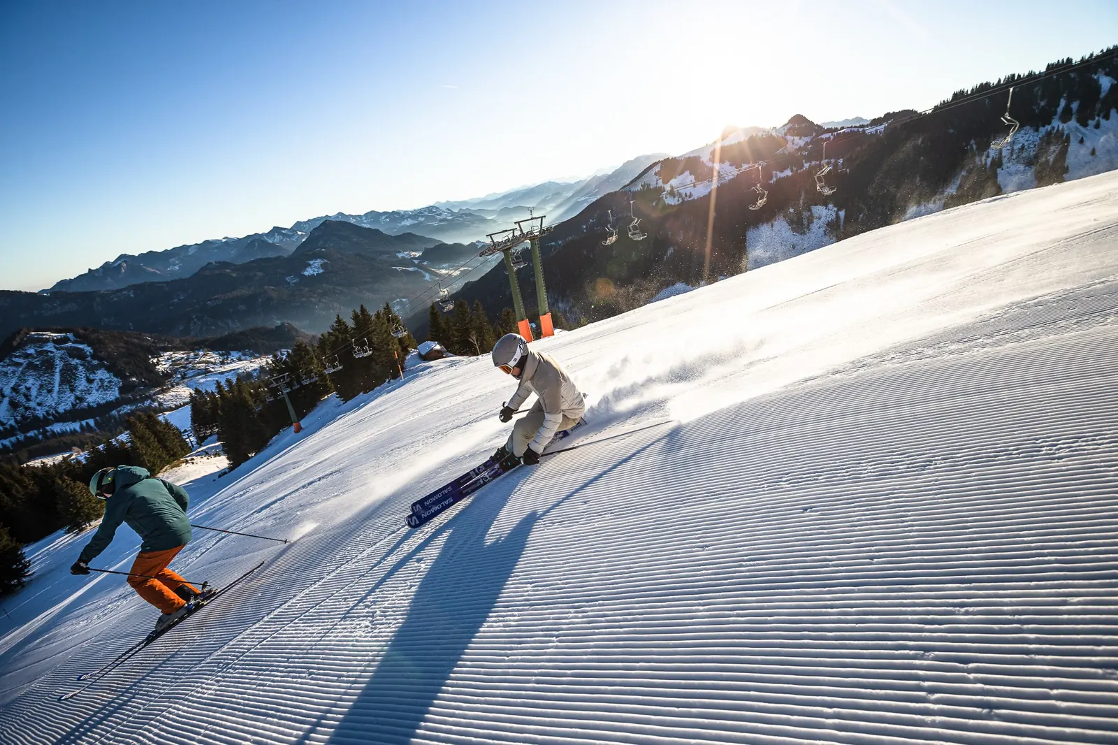 Zwei Skifahrer carven auf einer frisch präparierten Piste, im Hintergrund verschneite Berge und ein Sessellift