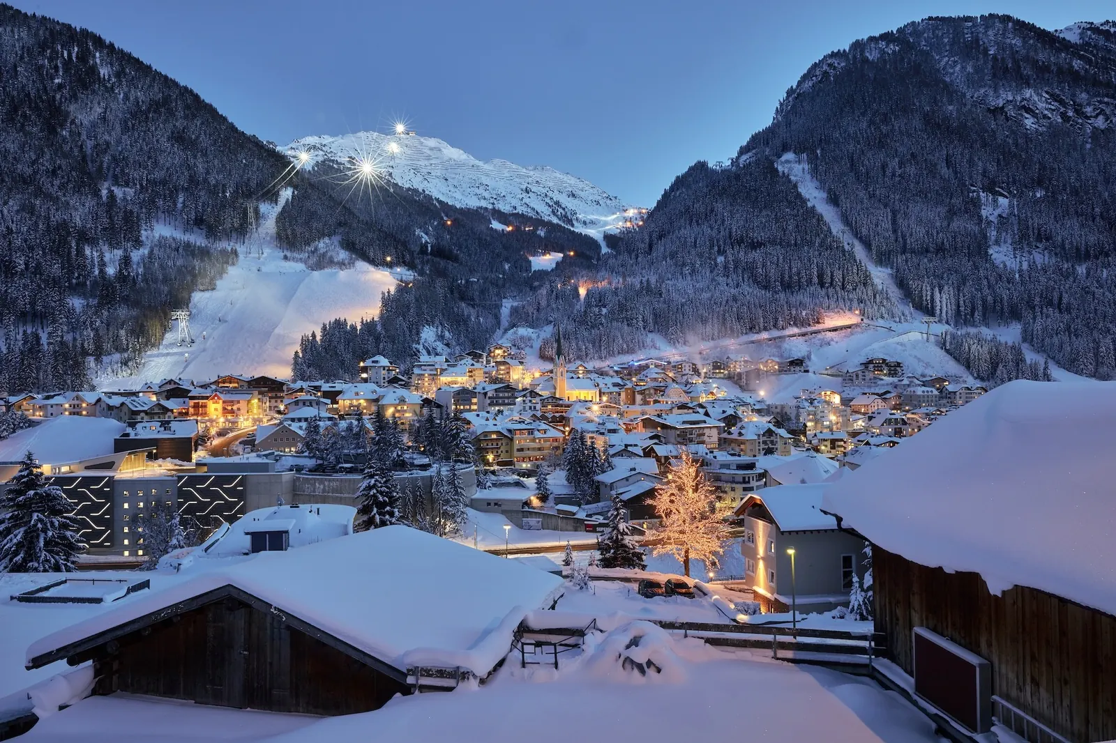 Verschneites Ischgl in der Abenddämmerung, dahinter beleuchtete Pisten am Hang