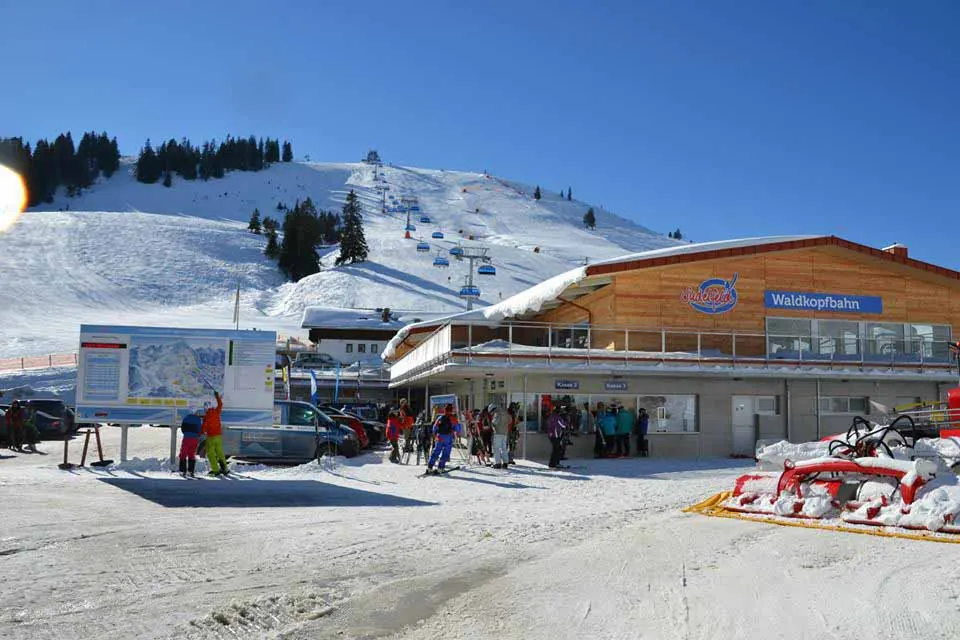 Talstation der Waldkopfbahn mit Servicegebäude, Pistenplan-Tafel und Skifahrern, dahinter der Sessellift am Hang