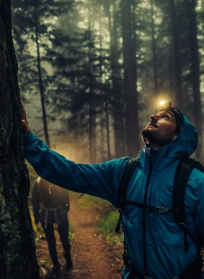 Wanderer in blauer The North Face Regenjacke mit Kapuze und Stirnlampe im nebligen Wald, vorhandenes Shopmotiv